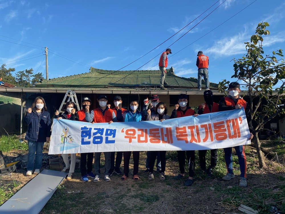 현경면 복지기동대 현수막을들고 사람들이 단체사진을 찍고 있고 그 뒤로 지붕위에 두사람이 올라가있다. 