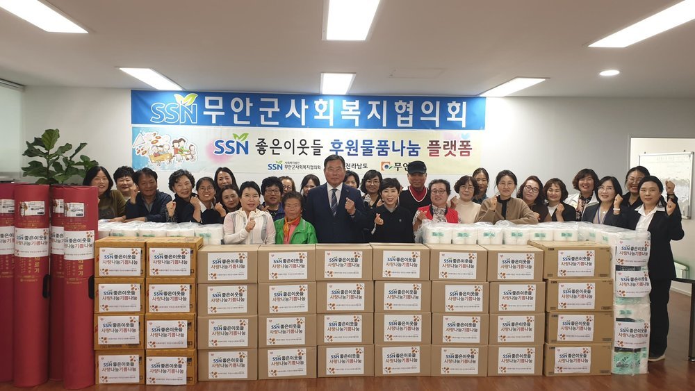 무안군사회복지협의회 SSN좋은이웃들 후원물품나눔 플랫폼이라고 쓰여진 현수막아래에 무안군사회복지협의회 회원들과 무안군수의 단체기념촬영 모습, 사람들 앞으로 기부물품 꾸러미들이 쌓여있다