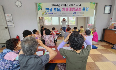 2024년 치매인식개선 사업의 한글호흡 치매예방교실 운영이라고 적힌 현수막을 중심으로 그 아래 서 이마를 마사지하는 모습의 강사를 따라하는 여러명의 수강생들의 모습