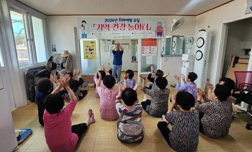 2024년 치매예방 교실의 기억 건강 놀이터라고 적힌 현수막 아래 서 있는 강사가 양손을 높이 들고 박수를 치고 있으며 수강생들이 따라하는 모습 