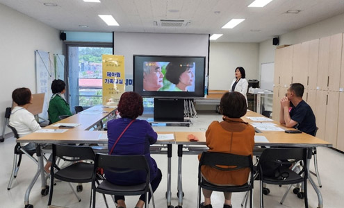 ㄷ자로 배치 된 테이블에 앉아 중앙에 있는 스탠드TV에 나오는 영상을 설명하는 강사를 바라보고 있는 수강생들의 모습