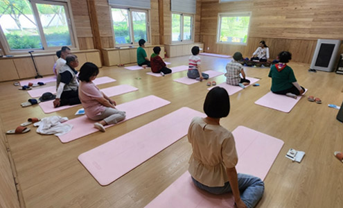 10개의 분홍색 요가매트를 5개씩 앞 뒤로 나누어 앉은 수강생들이 마주보고 앉은 강사를 바라보며 자세를 따라하는 모습